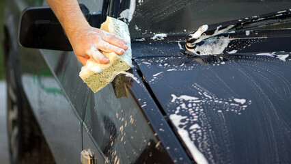 a hand with a washcloth and soap suds washes the hood of the car. Man washing car hood with washcloth and shampoo, auto covered with foam, car wash service. cleaning the black surface