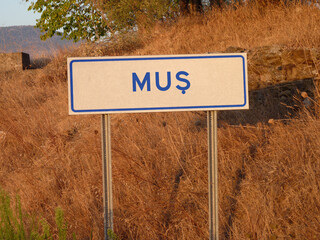 Blue 'Mus' Sign on a Metal Pole: A Bold Contrast Against the Natural Landscape of Brown Grass and Soft Shadows, Capturing a Serene Afternoon in the Heart of the City.