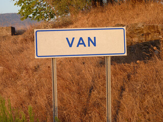 Blue 'Van' Sign on a Metal Pole: A Bold Contrast Against the Natural Landscape of Brown Grass and Soft Shadows, Capturing a Serene Afternoon in the Heart of the City.