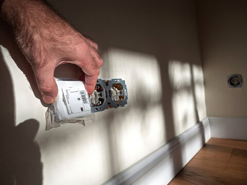 London, United Kingdom - Nov 14, 2020: Hand holding an electrical outlet cover near an exposed socket in a sunlit room, a home improvement or electrical process - Busch and Jaeger - Made in Germany