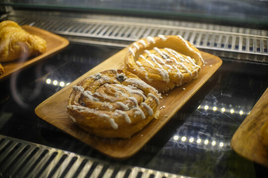 Two glazed Danish pastries displayed on a wooden tray inside a glass case. The golden pastries are drizzled with icing, showcasing their flaky layers and sweet filling.