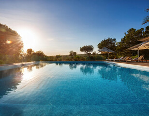 Obraz premium An clear swimming pool overlooking rolling hills at sunset, with pool chairs visible by the water's edge.