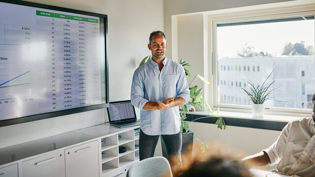Business Presentation in Modern Office with Data Analysis on Screen. A mature businessman, with a hint of gray in his hair, wearing a blue shirt, stands in front of his colleagues and speaks.