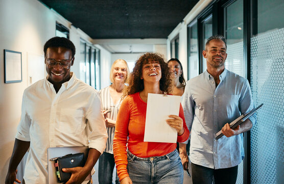 Diverse Team Walking Down Hallway in Modern Office Environment. They are all carrying some office supplies while smiling and laughing together.