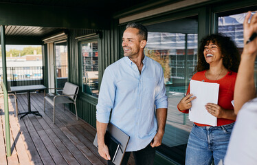 Smiling Business Colleagues Enjoying a Relaxed Outdoor Meeting on a Balcony. The businessman has a laptop under his arm, while the businesswoman holds some papers.