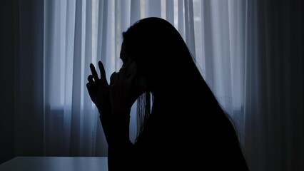 Silhouette of woman at table in front of window, talks on smartphone, screaming crying, drama and crisis, heartbroken expression. Human emotions and mental health.