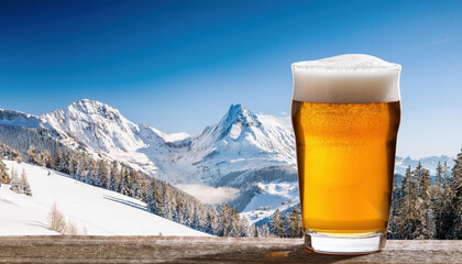 A glass of golden beer with foamy heads are positioned in snow with a winter mountain landscape copy space for your design