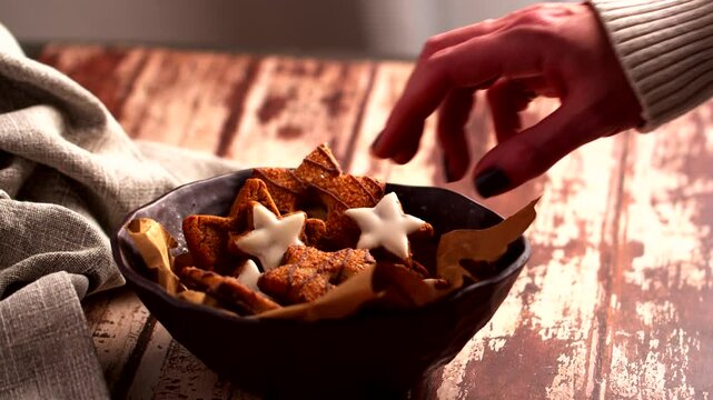 Female hand reaches for star-shaped cookies, some glazed with white icing, in a bowl lined with golden parchment paper. The rustic wooden table and soft fabric add a cozy, homemade feel to the scene.