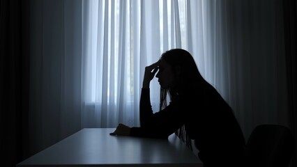 Silhouette of woman sitting at table in front of window, disappointed depressed expression, mental crisis, exhausted frustrated mood. Human emotions and mental health.