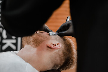Fototapeta premium Barber shop. Man with a beard a client in a barbershop hairdresser Barber on a shaving with a dangerous razor.Shaving process,face closeup.