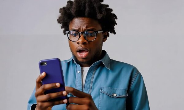 Homme afro avec lunettes regardant un smartphone bleu sur fond gris, portrait surpris en gros plan, r&eacute;action &eacute;tonn&eacute;e face &agrave; la technologie mobile, communication num&eacute;rique et usage du t&eacute;l&eacute;phone