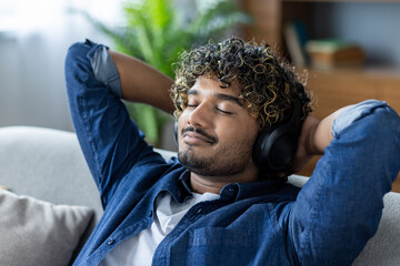 A young man is sitting on a couch, wearing headphones and enjoying music. He appears relaxed and content, embodying a peaceful, leisurely Saturday afternoon vibe.