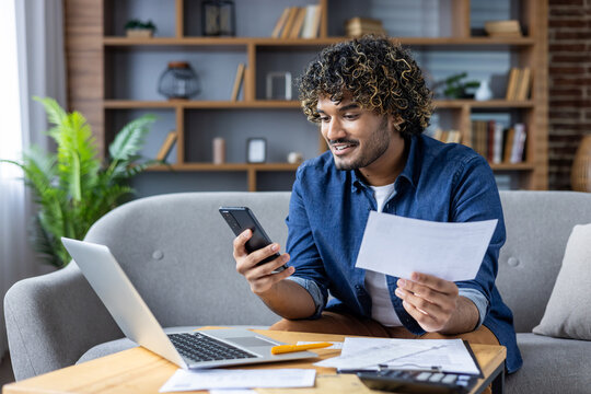A cheerful man sits on a sofa using his smartphone and laptop to manage his finances. He holds a paper document, illustrating the integration of technology in everyday tasks.