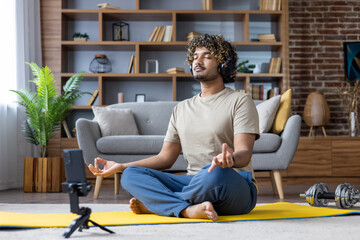 A man in a relaxed pose on a yoga mat meditates with headphones, surrounded by home decor. The...