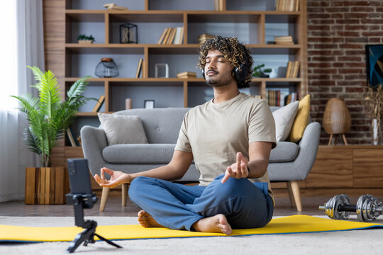 A man in a relaxed pose on a yoga mat meditates with headphones, surrounded by home decor. The phone beside him suggests a digital connection for guided meditation or music.