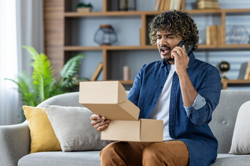 A man, visibly dissatisfied, communicates with an online store support service phone after receiving a damaged parcel. Seated on a sofa, his expression conveys frustration as he holds the open box.