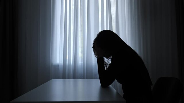 Silhouette of woman sitting at table in front of window. lays head on desk, depressed heartbroken expression, lonely and doubtful. Human emotions and mental health.