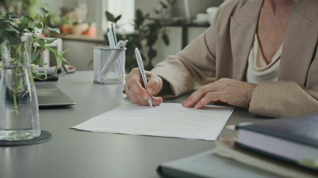Cropped shot of unrecognizable female entrepreneur taking pen and signing business document while sitting at modern desk, filled with office supplies and plants
