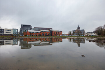 A placid body of water reflects modern apartment buildings and a church steeple on a cloudy day.  Urban landscape mirrored in still, muddy water.