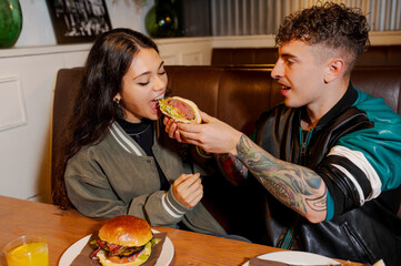 Young tattooed man feeding his girlfriend a burger at a restaurant table