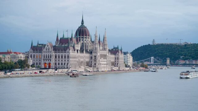 Hungarian Parliament in Budapest at sunset on cloudy day. Chain Bridge, Gell�rt Hill, Royal Palace in background. Boats gently drifting on the Danube River, capturing the city's charm at sunset.