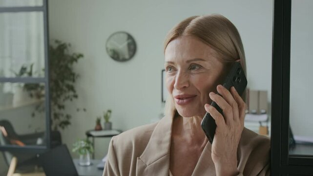 Medium close up with slowmo of mid-adult blonde Caucasian businesswoman talking on her smartphone while standing in modern office, filled with green plants and minimalist supplies and decor