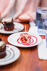 Elegant Dessert Presentation Featuring A Red Velvet Cake Pop And Decadent Chocolate Cupcakes On White Plates With Artistic Syrup Decoration And Glasses Of Water On A Wooden Table Setting