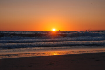The sun sets over the ocean at Venice Beach, Los Angeles, casting an orange glow. A boat silhouette is visible on the horizon, with waves lapping at the shore.