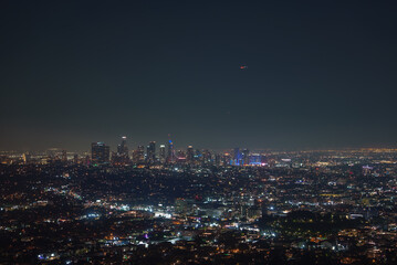 The Los Angeles skyline is illuminated at night, featuring the U.S. Bank Tower. City lights spread across the urban landscape, highlighting downtown.