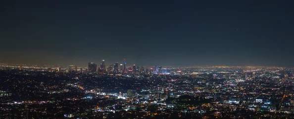 The Los Angeles skyline is illuminated at night, featuring iconic skyscrapers like the U.S. Bank Tower. City lights spread across the vast urban landscape. © Aerial Film Studio