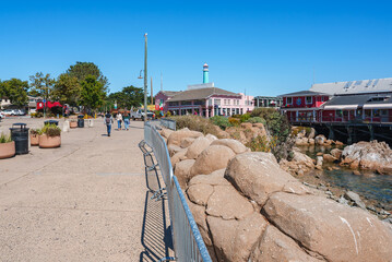 Cannery Row in Monterey, California features a rocky shoreline, colorful buildings, and a lighthouse like tower under a clear blue sky with people walking.