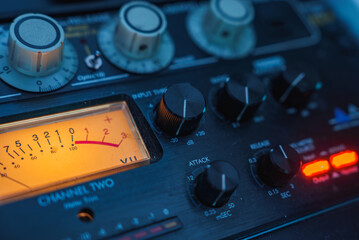 The image shows a detailed view of an audio mixing console, featuring various knobs, dials, and a glowing VU meter, indicating audio levels in a studio setting. © Aerial Film Studio