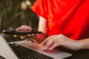 person in red shirt using smartphone while typing on laptop. scene captures blend of technology and...