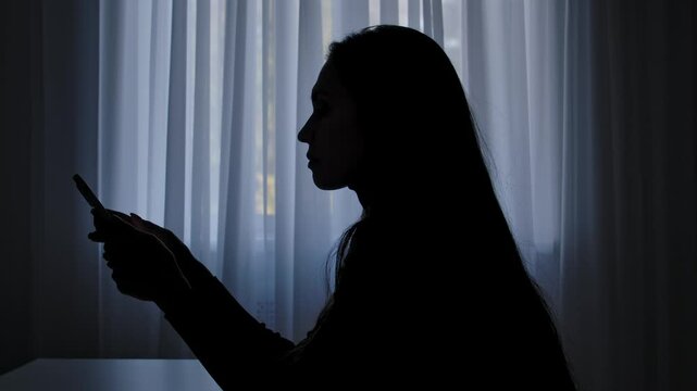 Silhouette of a woman at a table in front of a window, typing a message on a smartphone, drama and crisis, mental anguish. Human emotions and mental health.