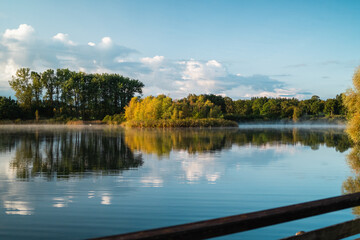 Fototapeta premium Serene Lake Landscape With Reflections of Lush Green Trees and Soft Morning Mist Under a Clear Blue Sky