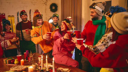 Festive holiday celebration with friends in ski-themed winter outfits gathered around wooden table,...