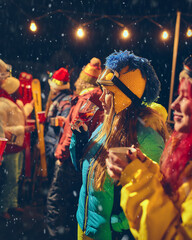 Friends in bright winter outfits celebrate outdoors with drinks, smiling and laughing as snow falls, creating festive holiday atmosphere. Concept of holidays, Christmas party. Ad