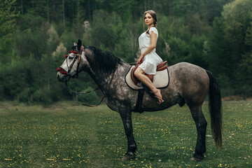 girl sitting on a horse. The girl is dressed in a white dress and barefoot. The horse is gray with...