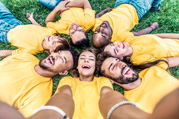 Happy best friends wearing yellow t shirt laughing at camera - Group of multiracial people taking selfie picture with smart mobile phone lying on the grass - Volunteers team smiling together outdoors