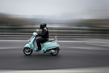 Blurred young woman on a green scooter, riding along a road to the left, with a panning effect.