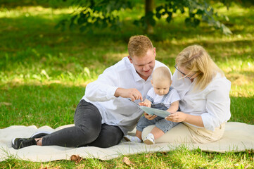 Fototapeta premium Parents with baby sitting on grass on summer day, reading interesting book to child, cute baby listening attentively. Children's education, communication, family vacation.
