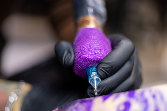 Close-up shot of a tattoo artist using a tattoo machine to create a detailed design on a client&rsquo;s arm, with a focus on the purple stencil and black gloves. Perfect for tattoo art, creativity, and life