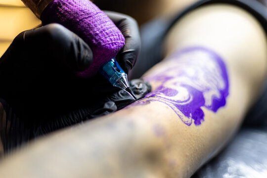 Close-up shot of a tattoo artist using a tattoo machine to create a detailed design on a client&rsquo;s arm, with a focus on the purple stencil and black gloves. Perfect for tattoo art, creativity, and life