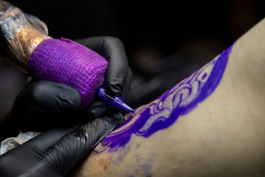 Close-up shot of a tattoo artist using a tattoo machine to create a detailed design on a client&rsquo;s arm, with a focus on the purple stencil and black gloves. Perfect for tattoo art, creativity, and life