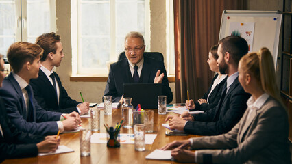 Senior business leader delivers presentation to group of young professionals during team meeting, with graphs and charts visible in background. Concept of business, corporate life, strategy planning
