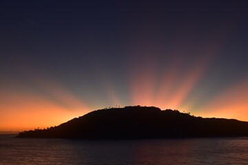 Stunning Pacific Ocean sunset with beams of colourful light shining up from the sea and islands in...