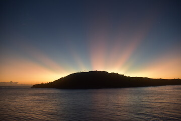 Stunning Pacific Ocean sunset with beams of colourful light shining up from the sea and islands in...