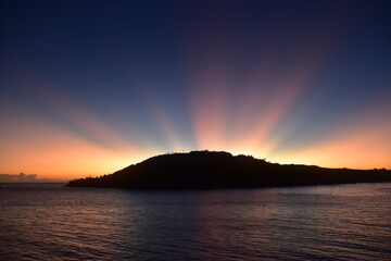 Stunning Pacific Ocean sunset with beams of colourful light shining up from the sea and islands in...