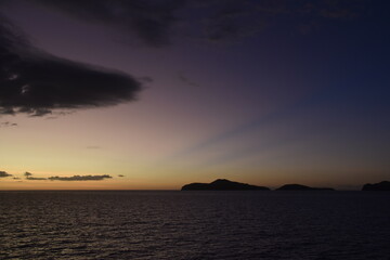 Stunning Pacific Ocean sunset with beams of colourful light shining up from the sea and islands in...