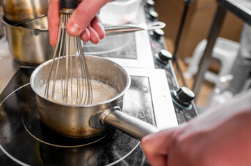 A close-up of hands whisking a mixture in a small saucepan on an electric stove. The focus is on the stainless steel pot and whisk, showcasing a cooking process in a kitchen setting.